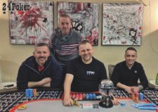 Ein Mann sitzt an einem Pokertisch und hält triumphierend Karten in der Hand, während er auf seinen Gewinn stolz lächelt. Um ihn herum stehen andere Spieler und beobachten das Spielgeschehen
