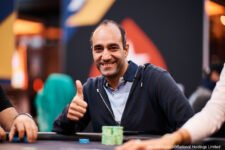 Ein Mann in einem dunkelblauen Anzug sitzt an einem Pokertisch und schaut konzentriert auf seine Karten. Er hat dunkles Haar und trägt eine Uhr am Handgelenk. Im Hintergrund