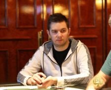 Ein Mann mit dunklen Haaren und einem schwarzen T-Shirt sitzt an einem Pokertisch und betrachtet konzentriert seine Karten. Im Hintergrund sind weitere Spieler und Karten auf dem Tisch zu sehen.