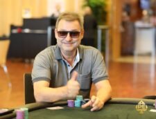 Ein Mann sitzt an einem Pokertisch, hält Karten in der Hand und schaut konzentriert auf den Tisch. Im Hintergrund sind andere Spieler und Casino-Chips zu sehen.