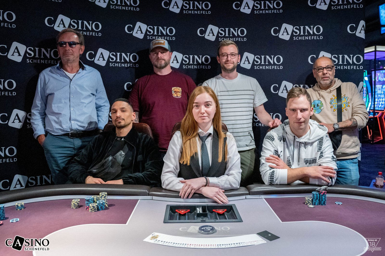 Ein Bild von einem Pokertisch während eines Turniers mit Spielern, die ihre Karten halten und Pokerchips vor sich stapeln. Im Hintergrund sind Zuschauer zu sehen.