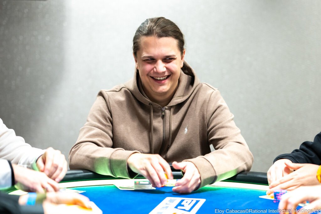 Ein Mann in einem dunkelblauen Anzug sitzt an einem Pokertisch und konzentriert sich auf sein Spiel. Er hat blonde Haare und trägt eine schwarze Sonnenbrille. Im Hintergrund sind andere Spieler