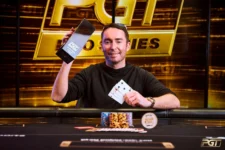 Ein Mann in einem schwarzen T-Shirt und einer Baseballmütze sitzt an einem Pokertisch und betrachtet konzentriert seine Karten. Im Hintergrund sind andere Spieler und Dealer zu sehen.