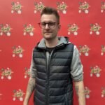 Ein Mann hält eine Hand mit Pokerkarten und schaut konzentriert auf den Tisch. Im Hintergrund sind weitere Spieler und Pokerchips zu sehen.