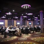 Ein Mann und eine Frau sitzen an einem Pokertisch und spielen eine Partie Poker. Der Mann hat einen Stapel Chips vor sich, während die Frau ihre Karten betrachtet. Im Hintergrund sind weitere Spieler und eine elegante