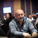 Ein Mann und eine Frau sitzen an einem Pokertisch in einem luxuriösen Casino. Der Mann hat Karten in der Hand und beide haben konzentrierte Gesichtsausdrücke. Im Hintergrund sind andere Spieler