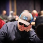 Ein Mann sitzt an einem Pokertisch und hält zwei Karten in der Hand, während er konzentriert auf seine Gegner schaut. Eine Reihe von Pokerchips liegt vor ihm auf dem Tisch.