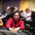 Ein Mann sitzt an einem Pokertisch und hält Karten in der Hand, während er konzentriert auf seine Gegner schaut. Die Szene spielt in einem eleganten Casino mit stilvoller Beleuchtung und