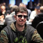 Ein Mann in einem dunkelblauen Anzug und weißem Hemd sitzt an einem Pokertisch und hält seine Karten in der Hand. Im Hintergrund sind weitere Spieler zu sehen, die gespannt auf die Karten