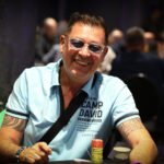 Ein Mann sitzt an einem Pokertisch und hält zwei Spielkarten in der Hand. Er hat einen konzentrierten Blick und trägt ein dunkles Hemd. Im Hintergrund sind weitere Spieler und Pokerchips zu sehen