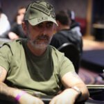 Ein Mann in einem dunkelblauen Anzug sitzt an einem Pokertisch und hält eine Hand mit Spielkarten. Er zeigt ein selbstbewusstes Lächeln, während er seinen Gegnern gegenüberblickt.