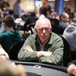 Ein Mann in einem dunkelblauen Anzug und einer weißen Krawatte sitzt an einem Pokertisch und hält seine Spielkarten in der Hand. Im Hintergrund sind andere Spieler und Chips auf dem Tisch zu sehen