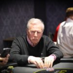 Ein junger Mann spielt Poker an einem eleganten Tisch in einem luxuriösen Casino. Er hält Karten in der Hand und betrachtet konzentriert die Gemeinschaftskarten auf dem Tisch. Andere