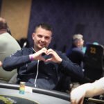 Ein Mann in einem dunkelblauen Anzug und einer roten Krawatte sitzt an einem Pokertisch und hält eine Hand mit Spielkarten. In der Mitte des Tisches liegen weitere Karten. Hinter ihm