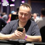 Ein Mann sitzt an einem Pokertisch, hält Spielkarten in der Hand und schaut konzentriert auf den Tisch. Im Hintergrund sind andere Spieler und Casino-Chips zu sehen.