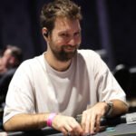 Ein Mann in einem Anzug und mit Sonnenbrille sitzt an einem Pokertisch und hält eine Hand mit Spielkarten. Andere Spieler sind im Hintergrund zu sehen.