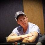 Ein Mann sitzt an einem Pokertisch und hält zwei Karten in der Hand. Er trägt einen Anzug und hat eine ernste Miene. Im Hintergrund sind weitere Spieler und Chips zu sehen.