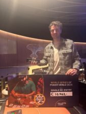 Ein Mann in einem dunklen Anzug und einer roten Krawatte sitzt an einem Pokertisch mit grüner Filzdecke. Er hält zwei Spielkarten in der Hand und schaut konzentriert auf seine
