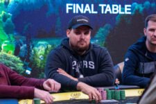 Ein Mann sitzt an einem Pokertisch und hält Pokerchips in der Hand. Er hat dunkle Haare und trägt ein schwarzes Hemd. Im Hintergrund sind weitere Spieler und Karten zu sehen.