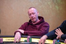 Ein Mann mit dunklen Haaren und Bart sitzt an einem Pokertisch und betrachtet konzentriert seine Karten. Er trägt ein dunkles Hemd und hat eine entschlossene Miene.