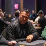 Ein Mann spielt Poker in einem Casino, umgeben von anderen Spielern und einer Dealerin. Er hat eine Hand aus Karten und Chips vor sich auf dem Tisch. Die Atmosphäre ist konzentriert und spannungsgel