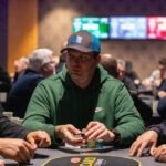 Ein Spieler sitzt an einem Pokertisch, hält Karten in der Hand und schaut konzentriert auf den Tisch. Um ihn herum sind Pokerchips und andere Spieler zu sehen.