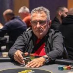 Ein Mann sitzt an einem Pokertisch und betrachtet konzentriert seine Karten, während im Hintergrund andere Spieler und Casino-Chips zu sehen sind.