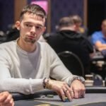 Ein Mann sitzt an einem Pokertisch und hält zwei Spielkarten in der Hand. Er trägt eine schwarze Kappe und hat eine ernste Miene. Im Hintergrund sind weitere Spieler und Pokerchips zu sehen.
