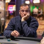 Ein Mann spielt Poker an einem eleganten Tisch in einem luxuriösen Casino. Er hält Karten in der Hand und hat einen konzentrierten Blick auf seine Gegner gerichtet. Die Atmosphäre ist gedämp