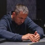 Ein Mann sitzt an einem Pokertisch und hält Karten in der Hand. Er trägt ein schwarzes Hemd und eine Sonnenbrille. Im Hintergrund sind weitere Spieler und Pokerchips zu sehen.