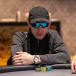 Ein Mann sitzt an einem Pokertisch und hält zwei Spielkarten in der Hand. Er wirkt konzentriert und umgeben von Pokerchips und anderen Spielern.