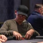 Ein Mann spielt Poker in einem eleganten Casino. Er hat eine ernste Miene und betrachtet seine Karten konzentriert, während er von anderen Spielern umgeben ist. Die Szene strahlt Spannung und Kon