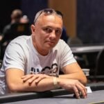 Ein Mann sitzt an einem Pokertisch und hält zwei Spielkarten in der Hand, während er intensiv auf die Gemeinschaftskarten auf dem Tisch schaut. Im Hintergrund sind weitere Spieler und Pokerchips zu