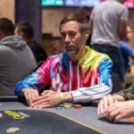 Ein Tisch mit einem Poker-Set, bestehend aus Karten, Chips und Getränken, umgeben von vier Personen, die konzentriert Poker spielen.