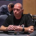Ein Mann und eine Frau sitzen an einem Pokertisch und spielen Poker. Beide haben Pokerchips vor sich liegen und halten Karten in der Hand. Der Mann trägt ein schwarzes Hemd und die Frau ein rotes Ob