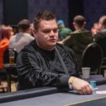 Ein Mann und eine Frau sitzen am Pokertisch, sie haben Karten in der Hand und schauen konzentriert auf den Tisch. Die Frau hat einen Stapel Chips vor sich liegen. Im Hintergrund sind weitere Spieler