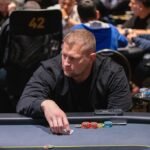 Ein Mann und eine Frau spielen Poker an einem eleganten Tisch in einem stilvollen Raum. Der Mann hält Karten in der Hand, während die Frau konzentriert auf ihre Karten schaut. In der Mitte des T