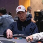 Ein Mann in einem dunkelblauen Anzug und einer roten Krawatte sitzt an einem Pokertisch mit Chips und Karten. Er hält zwei Spielkarten in der Hand und betrachtet sie konzentriert.