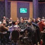 Ein Mann in einem dunkelblauen Anzug und weißem Hemd sitzt an einem Pokertisch und hält eine Hand mit Spielkarten. In der Mitte des Tisches liegen weitere Karten und Jetons. Im H