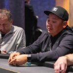 Ein Mann in einem dunklen Anzug und einer schwarzen Krawatte sitzt an einem Pokertisch und betrachtet konzentriert seine Karten. Im Hintergrund sind andere Pokerspieler und Spielchips zu sehen
