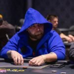 Ein Mann sitzt an einem Pokertisch und hält zwei Karten in der Hand, während er konzentriert auf die Gemeinschaftskarten schaut. Andere Spieler sind im Hintergrund zu sehen.