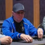 Ein Mann und eine Frau sitzen an einem Pokertisch und spielen Karten. Der Mann hat eine Hand mit Chips vor sich, während die Frau ihre Karten betrachtet. Im Hintergrund sind weitere Spieler und eine Glühbirne