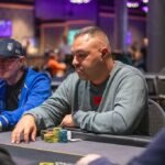 Ein Mann mit einem roten Hemd und einer Sonnenbrille sitzt an einem Pokertisch mit Karten und Pokerchips. Er hat eine ernste Miene und scheint konzentriert zu sein.