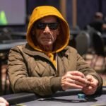 Ein Mann sitzt an einem Pokertisch und hält zwei Spielkarten in der Hand. Er wirkt konzentriert und umgeben von anderen Pokerspielern.