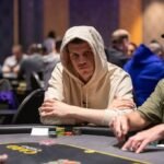 Ein Mann sitzt an einem Pokertisch, umgeben von anderen Spielern. Er hat eine Hand aus Spielkarten in der Luft und schaut konzentriert auf den Tisch. Die Atmosphäre ist angespannt und