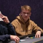 Ein Mann sitzt an einem Pokertisch und hält Karten in der Hand, während er konzentriert auf seine Gegner schaut. Im Hintergrund sind Pokerchips und Getränke zu sehen.
