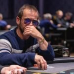 Ein Mann und eine Frau sitzen an einem Pokertisch und spielen Karten. Der Mann hat eine Hand mit Assen, während die Frau ernsthaft ihre Karten betrachtet. Im Hintergrund sind Pokerchips und Getr