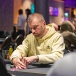 Ein Mann sitzt an einem Pokertisch und hält zwei Spielkarten in der Hand, während er konzentriert auf seine Gegner schaut. Die Chips auf dem Tisch sind bunt und stapeln sich hoch.