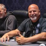 Ein Spieler hält zwei Spielkarten in der Hand und betrachtet sie konzentriert. Im Hintergrund sind Pokerchips und eine grüne Pokertischdecke zu sehen.