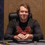 Ein Mann sitzt lächelnd an einem Pokertisch in einem Casino und hält zwei Karten in der Hand, während er auf seine Gegner schaut. Die Spannung und Konzentration ist in der Luft spürbar.